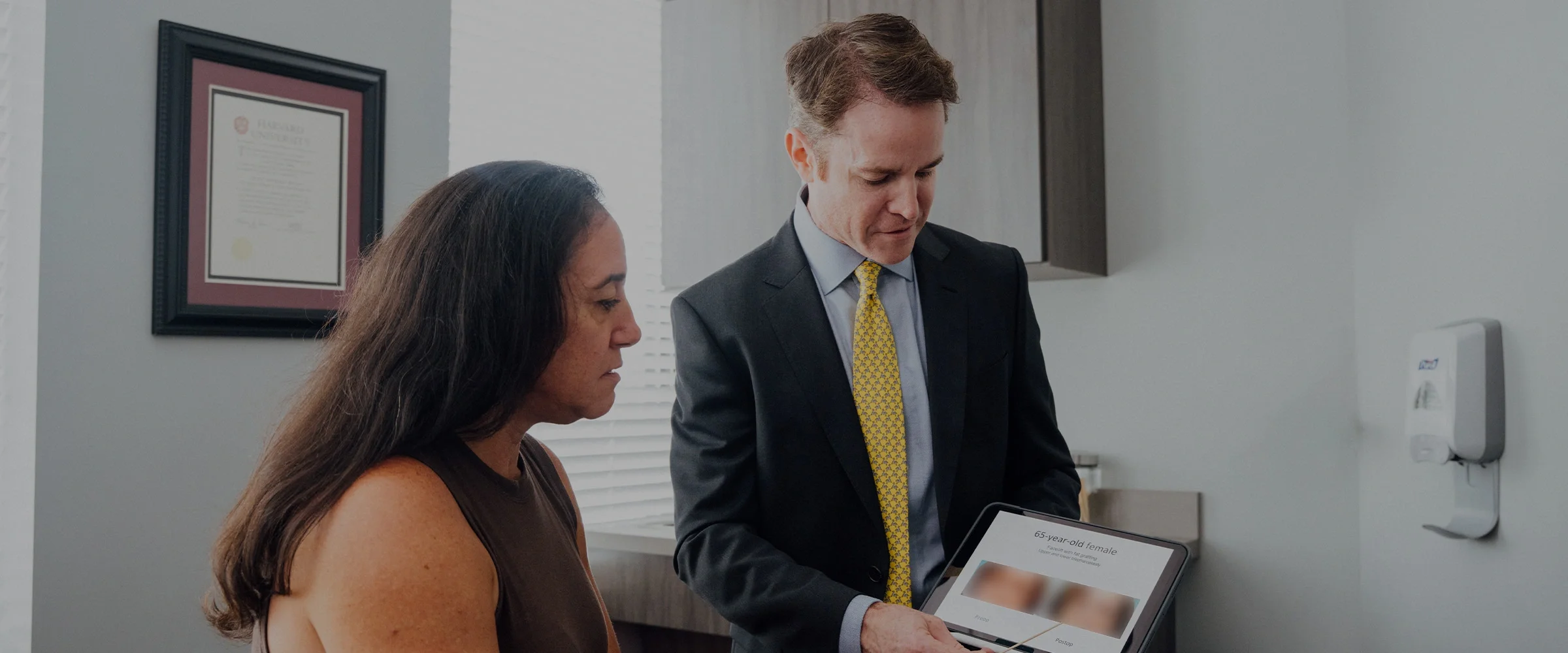 A physician, Dr. Henry Wilson, wearing a dark suit and yellow tie, shows a tablet to a seated female patient with long dark hair in an examination room. The tablet screen displays before-and-after photos of a woman's face. The patient is looking intently at the tablet. A framed certificate hangs on the wall behind the patient. - Face Procedures in Huntersville, NC