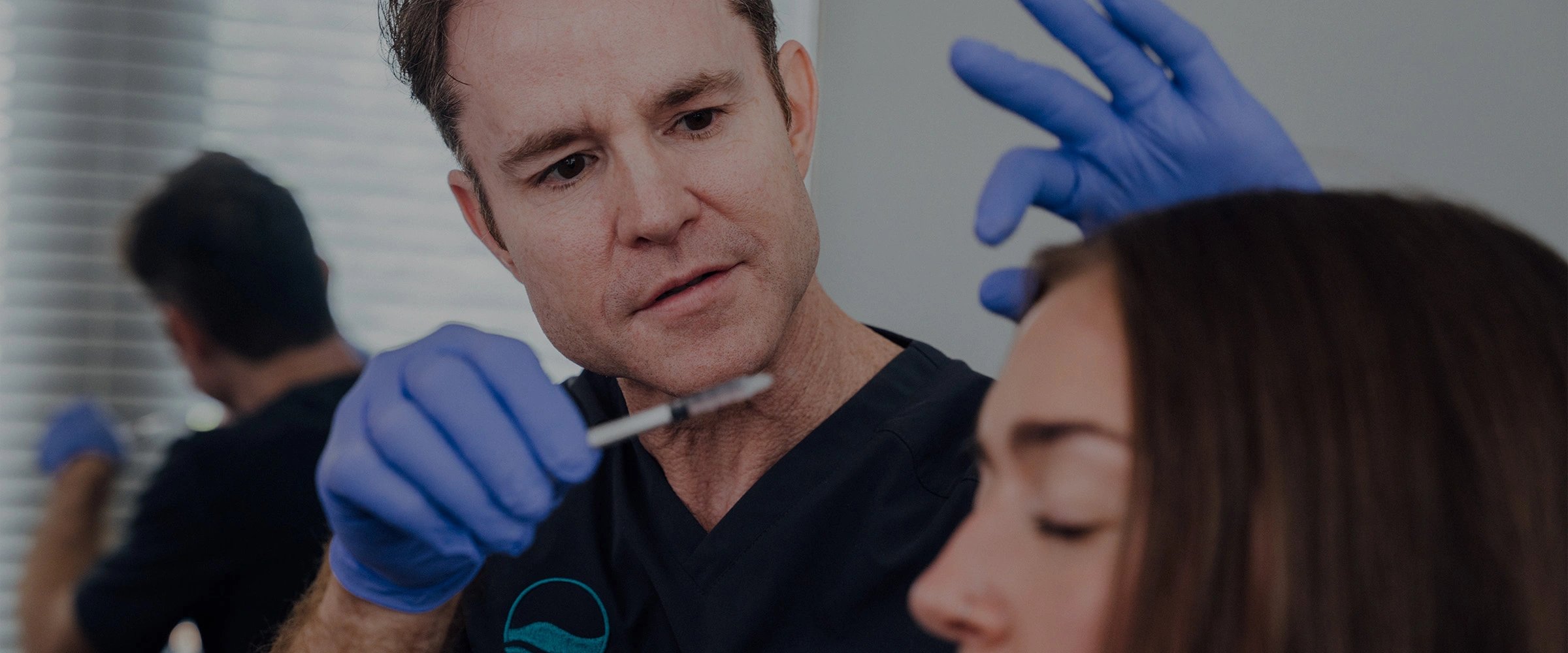 Dr. Henry Wilson, wearing a dark scrub top and blue gloves, is preparing to give an injection to a female patient, whose eyes are closed. He is holding a syringe close to the patient's face with one hand, and his other hand is near her temple. - Non-Surgical in Huntersville, NC