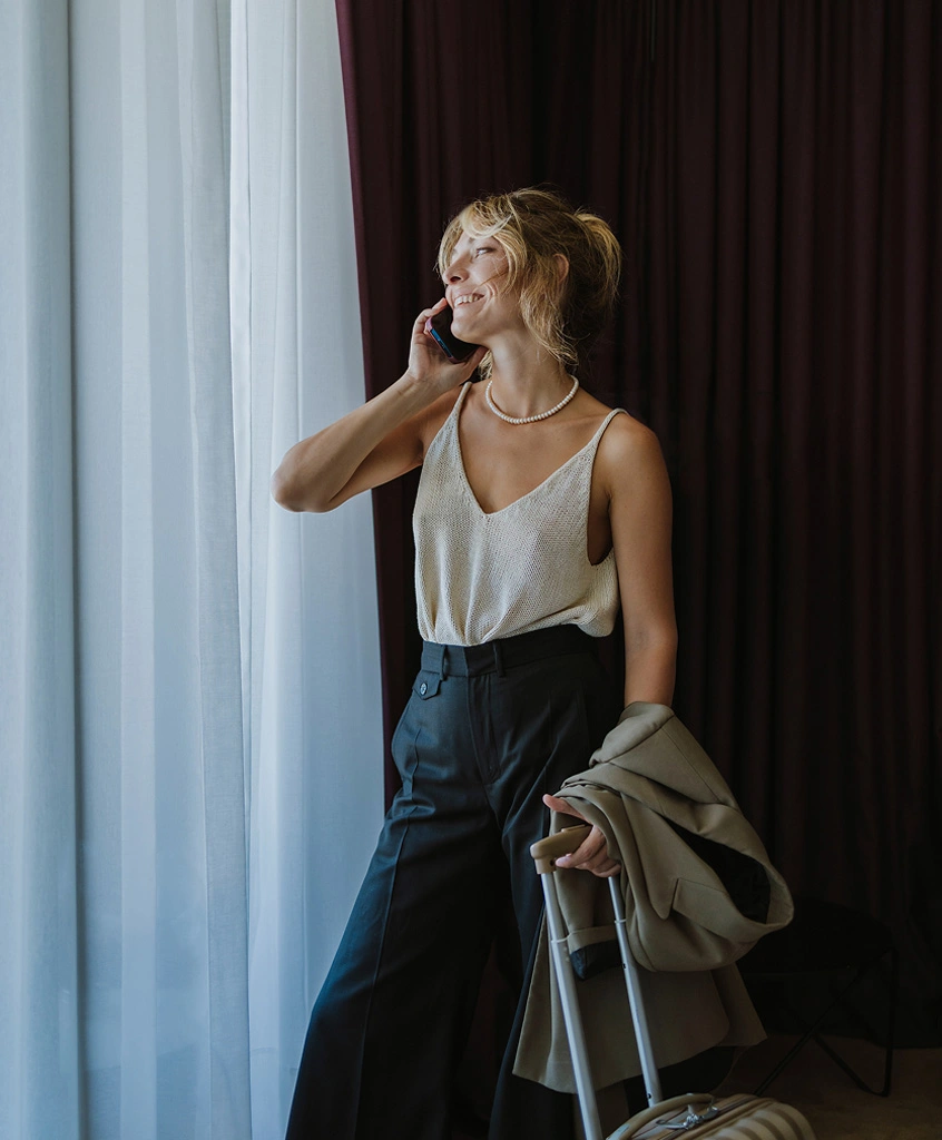 A smiling woman is standing by a window with white sheer curtains and dark drapes, talking on a cell phone while wearing a cream-colored tank top and black wide-leg pants. She is holding a jacket and resting her hand on a suitcase. - Traveling in Huntersville, NC