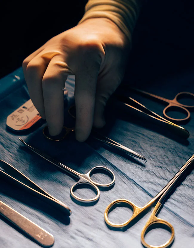 A gloved hand reaches for a piece of surgical equipment from a tray of metal tools, including scissors and tweezers. - Skin Cancer Screening Excision with Reconstruction in Huntersville, NC