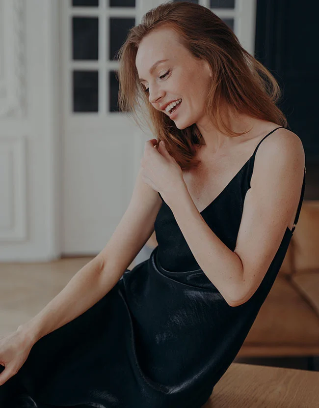 A light-skinned woman with shoulder-length reddish-brown hair is seated, smiling warmly and looking slightly to her left. She wears a dark, likely black, slip dress or top with spaghetti straps. Her joyful expression is complemented by soft, natural lighting. The background shows a light-colored wall and windows, creating a cozy, inviting atmosphere. - Arm Lift in Huntersville, NC