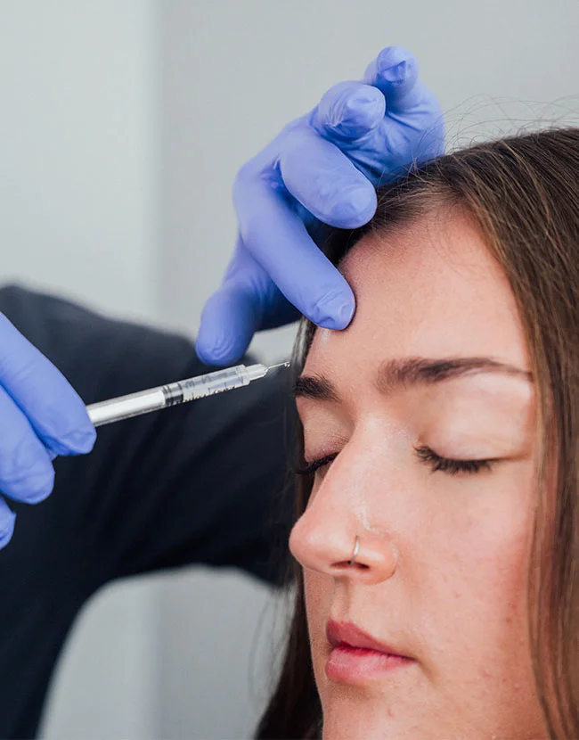 A medical professional wearing blue gloves is injecting a needle, likely for Botox or a wrinkle relaxer, into the forehead of a female patient with her eyes closed. - Botox and Wrinkle Relaxers in Huntersville, NC