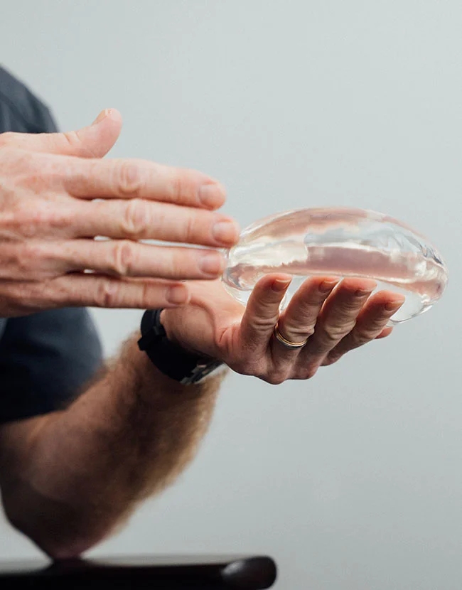 A pair of hands holding and gesturing over a clear, teardrop-shaped silicone breast implant. - Breast Augmentation in Huntersville, NC
