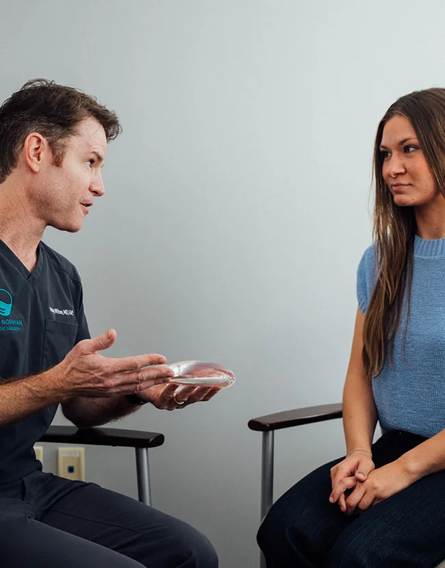 Close up of Dr. Henry Wilson, wearing dark blue scrubs, sits facing a young female patient in a light blue sweater. The doctor is holding and discussing a breast implant with the patient, who is looking down and listening. - Breast Implant Revision Surgery in Huntersville, NC