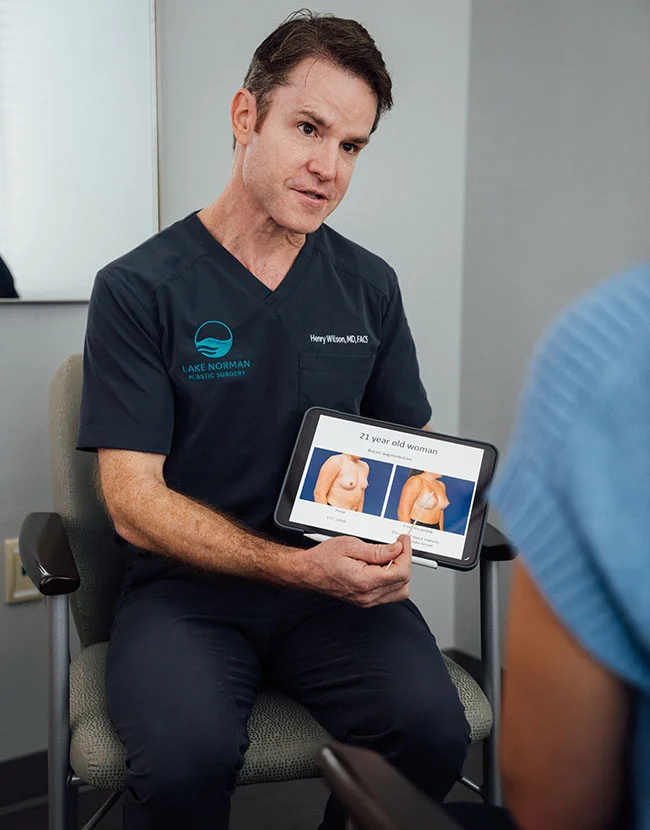 A male medical professional, Dr. Henry Wilson, wearing a dark scrub top, is sitting and showing a patient a tablet that displays before-and-after images related to breast surgery. - Breast Lift with Augmentation in Huntersville, NC