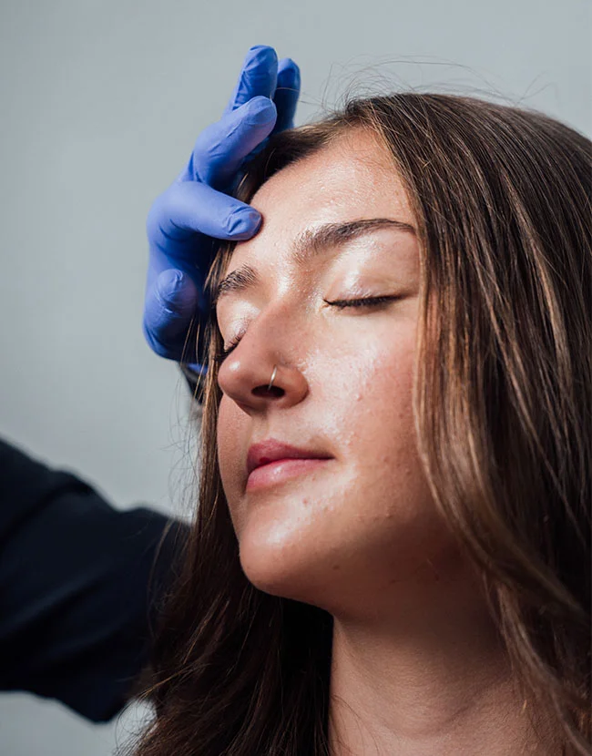 A medical professional wearing a blue glove gently places a hand on the forehead of a young woman with her eyes closed. - Browlift in Huntersville, NC