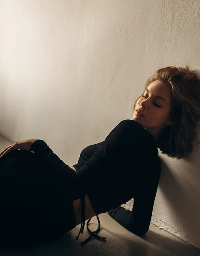 A young woman in a black, long-sleeve crop top is lying on a light-colored floor, leaning against a white wall with her eyes closed. - Buttock Lift in Huntersville, NC