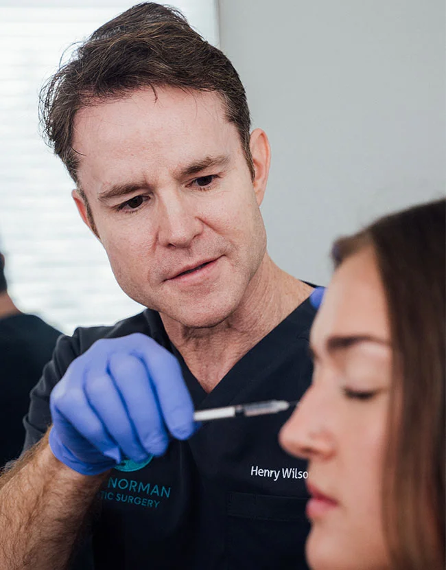 A male medical professional, Dr. Henry Wilson, wearing a dark scrub top and blue gloves, is administering an injection, likely a dermal filler, near the nose and cheek of a female patient. - Dermal Fillers in Huntersville, NC