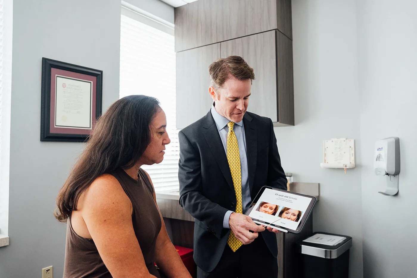 A physician, Dr. Henry Wilson, wearing a dark suit and yellow tie, shows a tablet to a seated female patient with long dark hair in an examination room. The tablet screen displays before-and-after photos of a woman's face. The patient is looking intently at the tablet. A framed certificate hangs on the wall behind the patient.