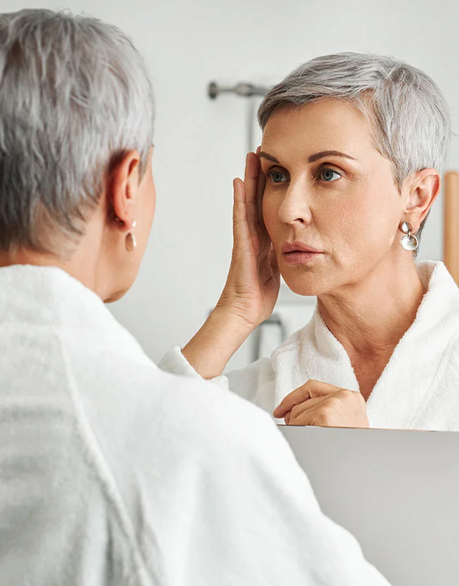 A woman with short, gray hair is looking at her reflection in a mirror. She is wearing a white bathrobe. She is touching her face, and her expression appears somewhat concerned or pensive. The lighting in the bathroom is bright and neutral. The background is a light gray/white bathroom wall. The reflection in the mirror shows a similar-aged woman with a similar expression. - Eyelid Surgery in Huntersville, NC