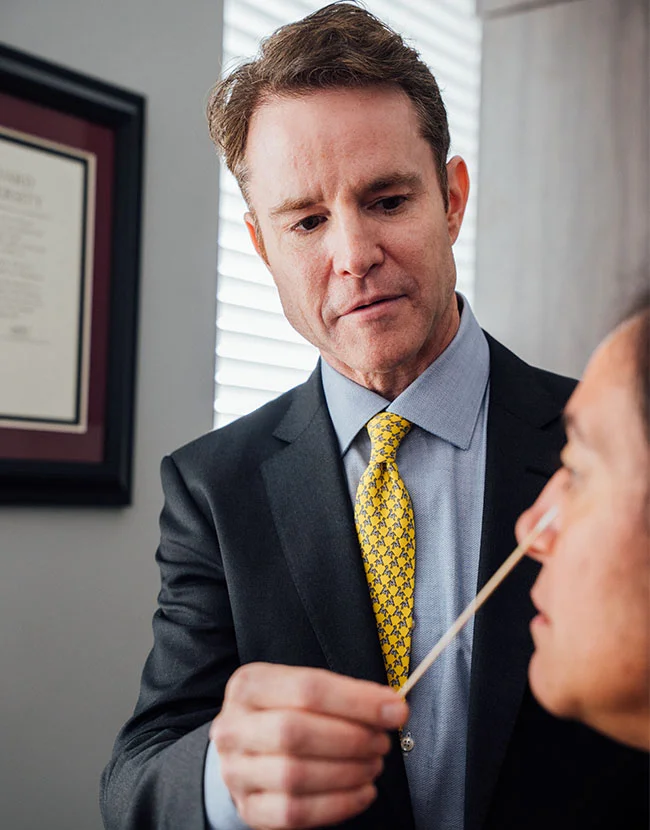 A male medical professional, Dr. Henry Wilson, wearing a suit and yellow tie, is using a small cotton swab to gently touch the nose of a patient. - Facial Fat Grafting in Huntersville, NC