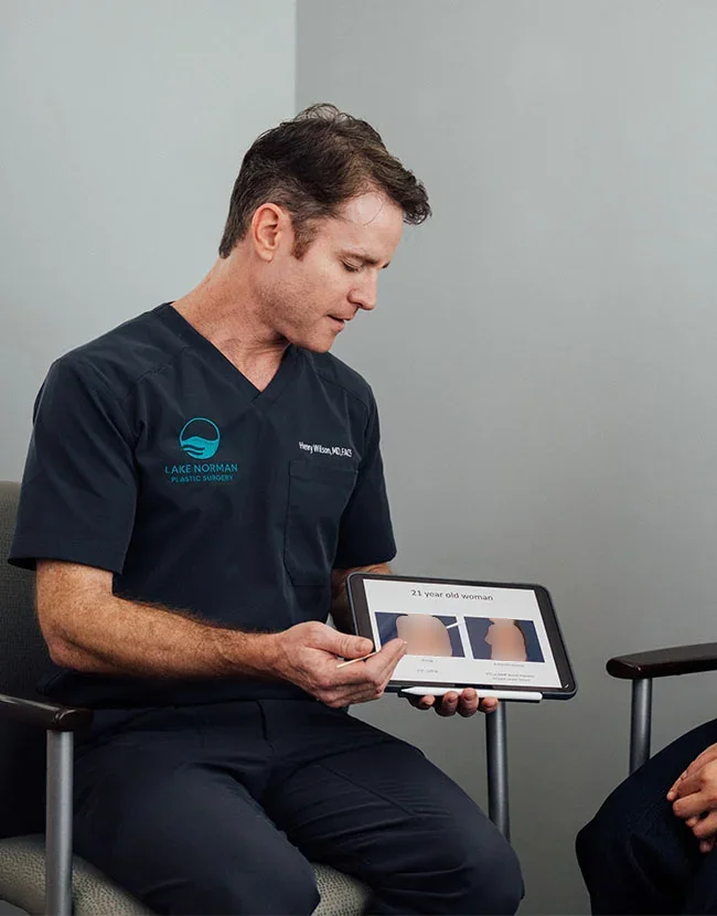 A male medical professional, Dr. Henry Wilson, wearing a dark scrub top, is sitting and showing a female patient a tablet displaying before-and-after images related to breast augmentation. - Fat Transfer Breast Augmentation in Huntersville, NC