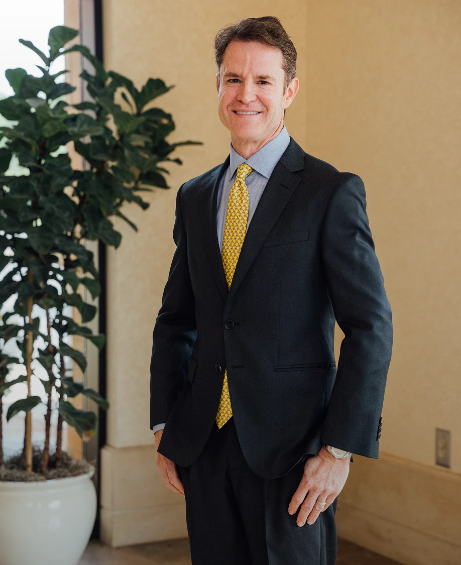 A portrait shot of Dr. Henry Wilson, wearing a dark suit and yellow patterned tie, smiles while standing indoors next to a large potted plant.