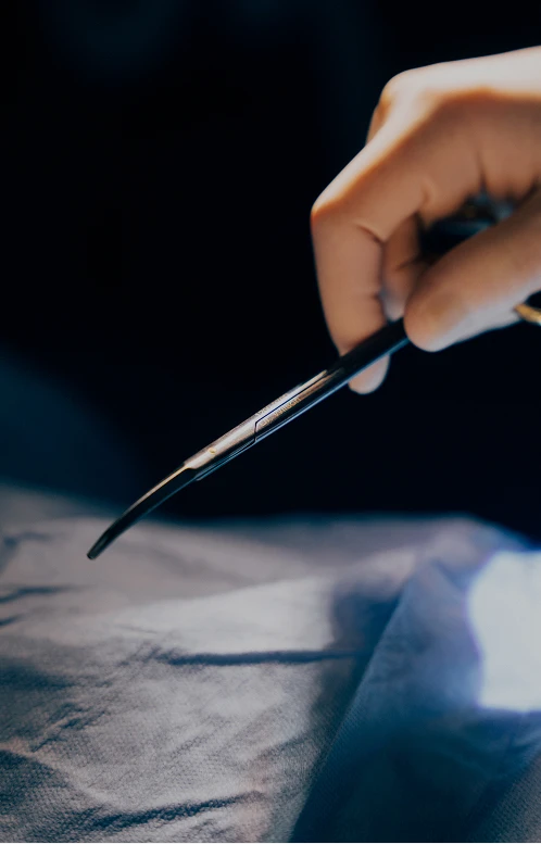 A close-up, dimly lit image of a hand holding a pair of curved surgical scissors or forceps over a dark blue surgical drape.