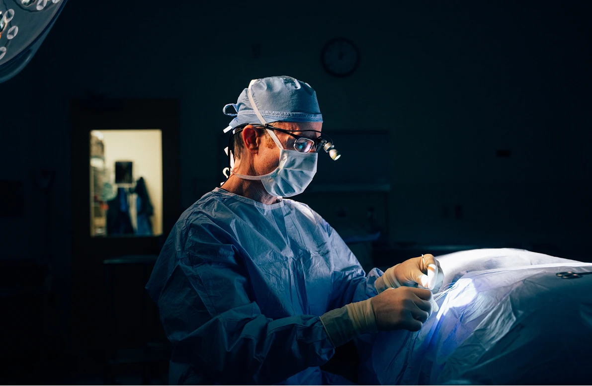 Dr. Henry Wilson, a surgeon, is shown in a dimly lit operating room, illuminated by a surgical light. He is wearing blue surgical scrubs, a cap, a face mask, safety goggles, and a headlamp. He is looking down and working over a patient draped in a white sheet. A clock is visible on the dark wall behind him.