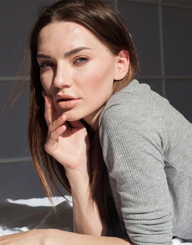 A young woman with long brown hair is resting her chin on her hand, looking directly at the camera while wearing a grey ribbed shirt. - Lip Fillers in Huntersville, NC