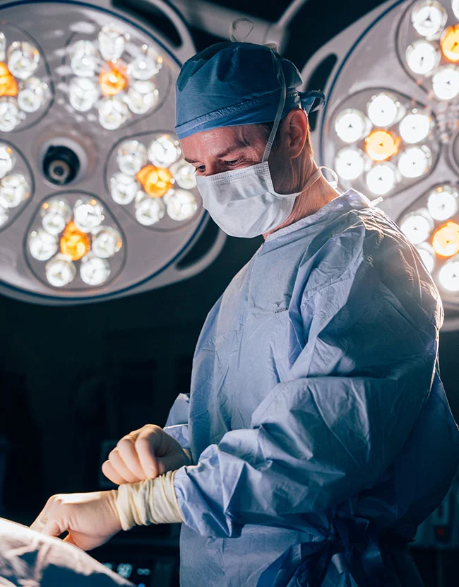 A male surgeon, Dr. Henry Wilson, wearing a blue scrub suit, surgical cap, and mask, is looking down and concentrating while performing a procedure under bright operating room lights. - Liposuction in Huntersville, NC