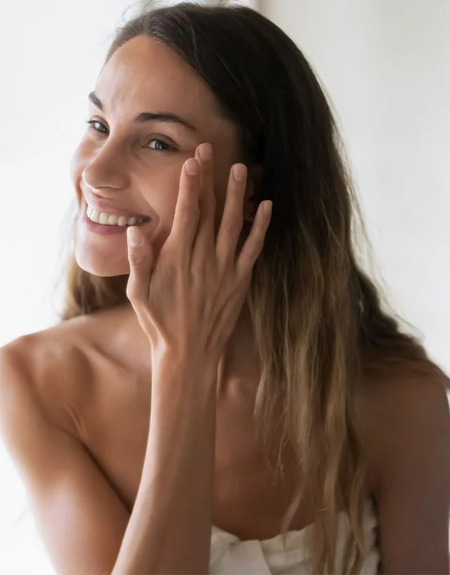 A smiling young woman is touching her cheek with her fingers, suggesting she is applying a skincare product. - Medical Grade Skincare Products in Huntersville, NC