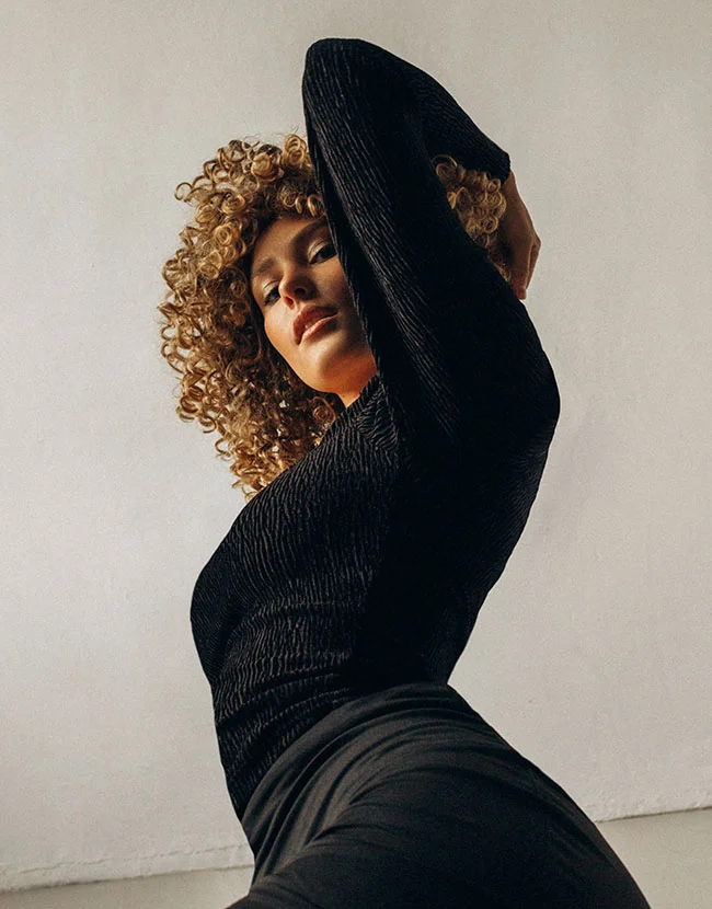 A woman with voluminous blonde curly hair is looking down at the camera, wearing a textured black long-sleeve top, with one arm raised over her head. - Mommy Makeover in Huntersville, NC