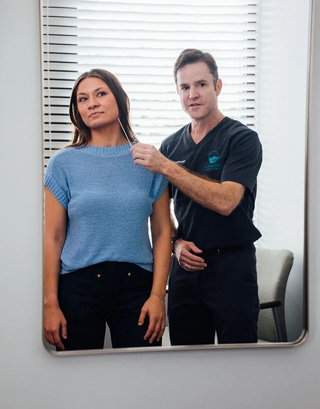 A female patient in a blue top and a male medical professional, Dr. Henry Wilson, wearing a dark scrub top, are reflected in a mirror. Dr. Wilson is holding a small stick, possibly a measuring or marking tool, near the patient's jawline. - Neck Lift in Huntersville, NC