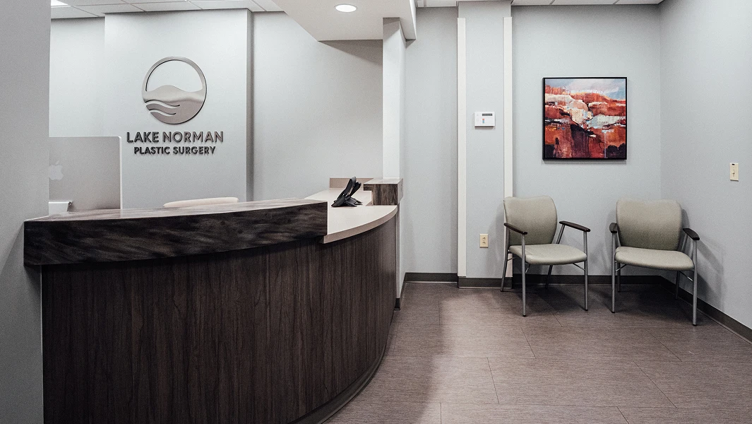 A photograph of the Lake Norman Plastic Surgery office reception area, showing a curved wood-paneled desk, the clinic's silver logo on the wall, two waiting chairs, and a piece of abstract art.