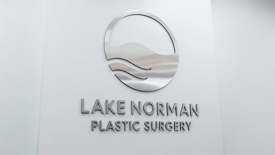 A close-up image of the Lake Norman Plastic Surgery logo and name mounted in silver metal on a white wall. The logo features a circle with waves inside, representing water.