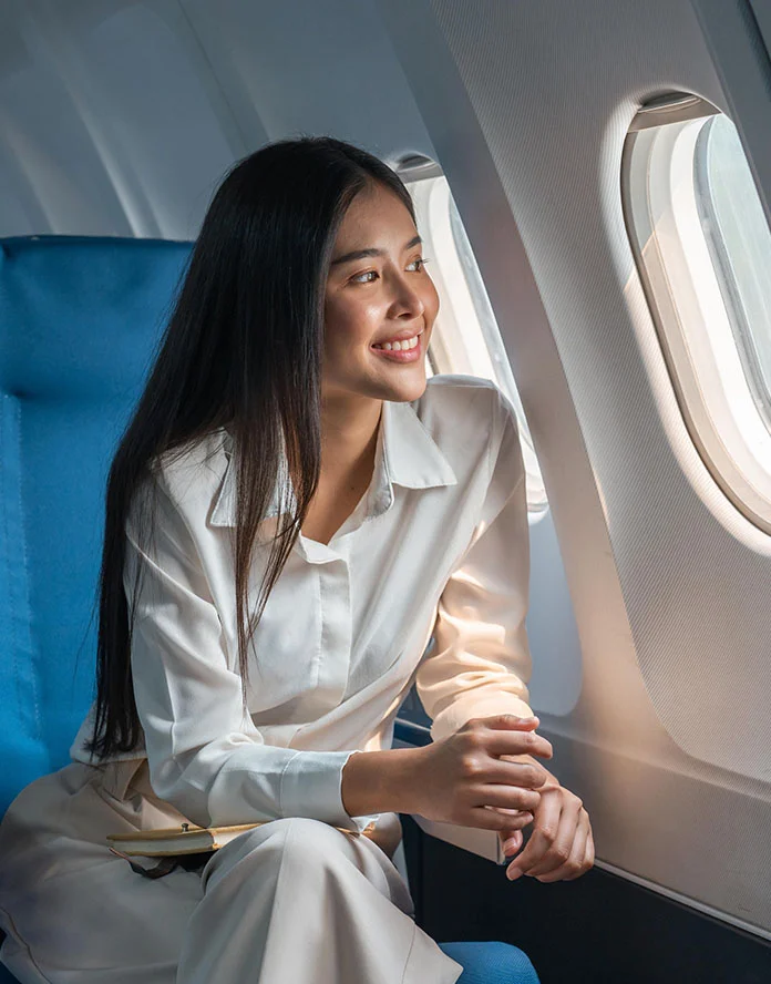 A smiling woman with long dark hair is seated in a blue airplane seat next to a window, looking out while wearing a white button-down shirt. She is resting her hands on her lap. - Out of Town in Huntersville, NC