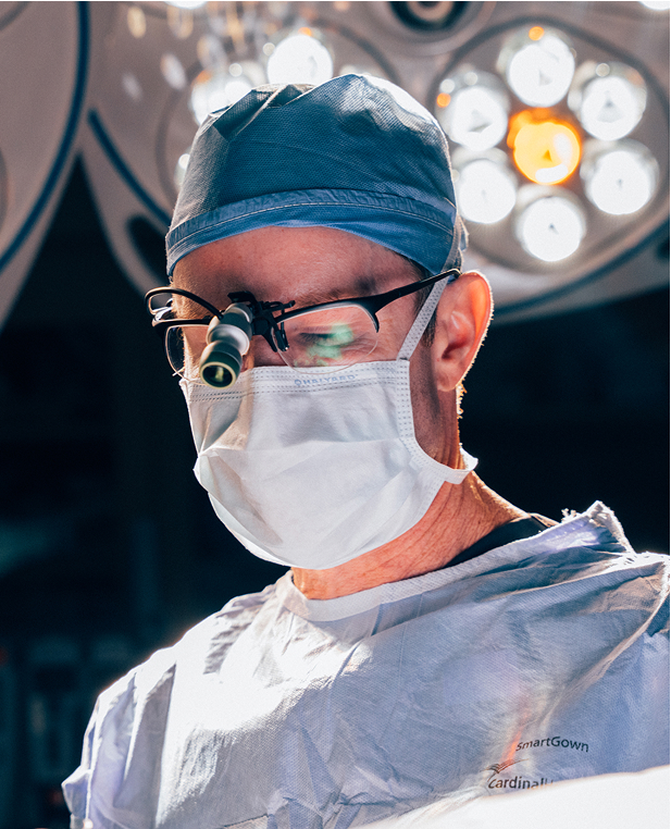 Close up shot of Dr. Henry Wilson outdoors, wearing protective scrubs, mask and hair cap. He wears glasses and a magnifying glass emphasizing his precision.