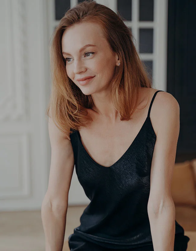 A fair-skinned woman with auburn hair is smiling and looking to the side while wearing a black satin camisole. - Scar Revision in Huntersville, NC