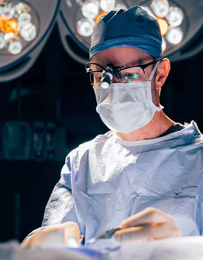 Close up shot of Dr. Henry Wilson outdoors, wearing protective scrubs, mask and hair cap. He wears glasses and a magnifying glass emphasizing his precision. - Tummy Tuck in Huntersville, NC